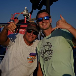 Two men posing with thumbs up on a boat, wearing casual t-shirts and sunglasses.
