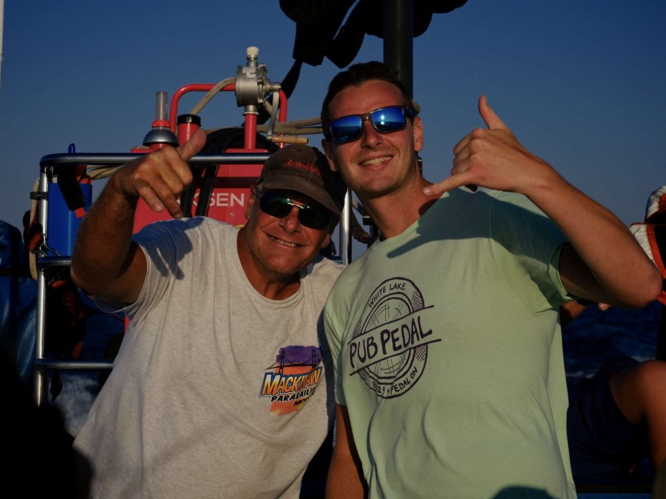Two men posing with thumbs up on a boat, wearing casual t-shirts and sunglasses.