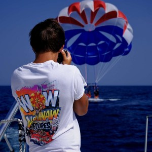 Person on boat photographing parasailers on ocean under a clear blue sky.