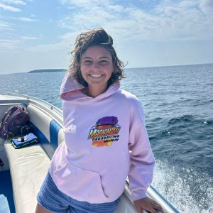 Person in pink hoodie smiling on a boat with ocean in the background.