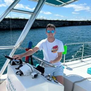 Person in sunglasses steering a boat on a sunny day with blue water and sky.