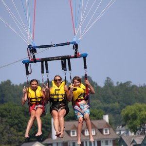 Three people parasailing with yellow life jackets over water with trees and buildings in background.