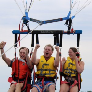 Three people in life vests parasailing, smiling and cheering in the air.