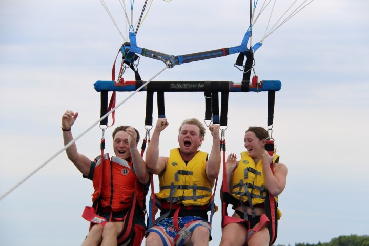 Three people in life vests parasailing, smiling and cheering in the air.