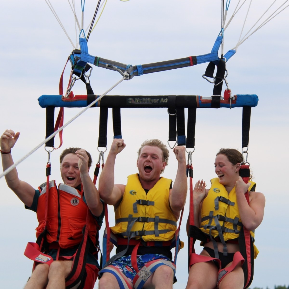 Three people in life vests parasailing, smiling and cheering in the air.