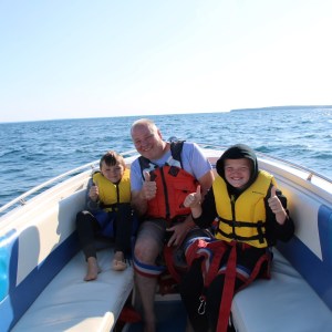 Three people in life jackets on a speedboat, smiling and giving thumbs up on a sunny day.