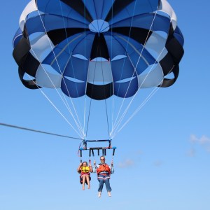 Two people parasailing under a blue parachute against a clear sky.