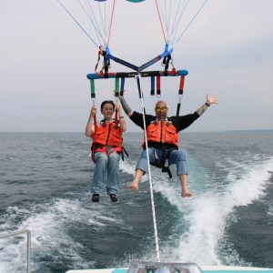 Two people parasailing over the ocean, wearing life jackets, with a boat trailing below.