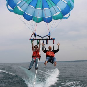 Two people parasailing with blue parachute over water, wearing life vests.